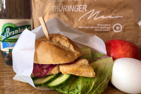 Ein schmackhaft belegtes Br&ouml;tchen, ein Ei, ein Apfel und ein Getr&auml;nk stehen vor einer Verpackung mit der Aufschrift Genusspaket Th&uuml;ringer Meer