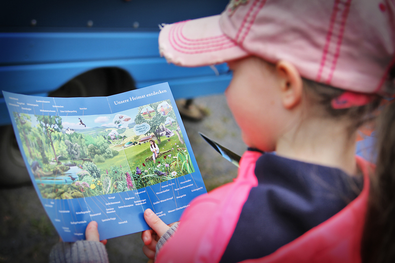 Ein M&auml;dchen h&auml;lt einen Flyer mit Wimmelbild vor sich ge&ouml;ffnet und guckt hinein.